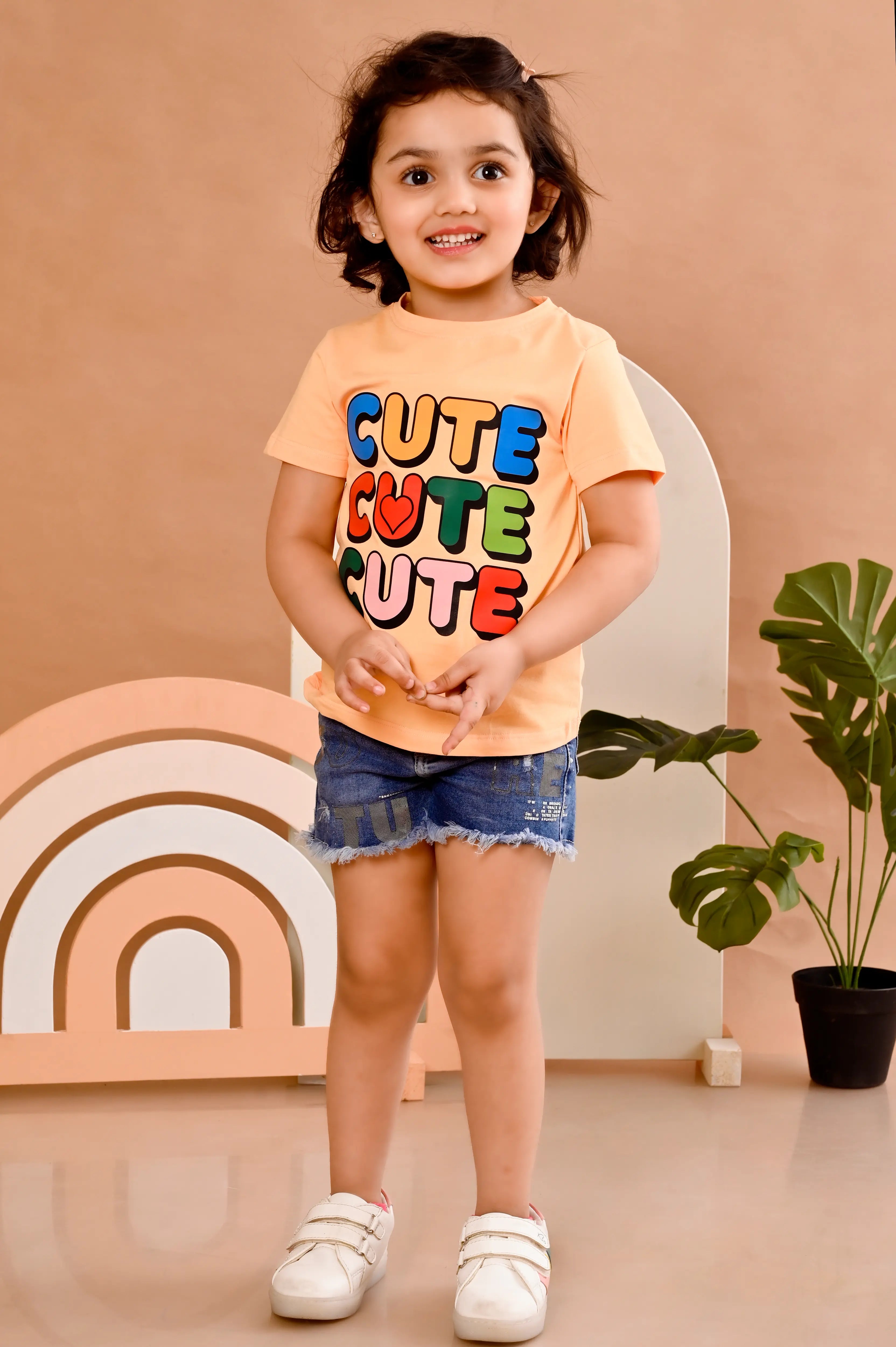Child wearing a 'CUTE CUTE CUTE' shirt with colorful text, standing in front of a decorative arch and plant.