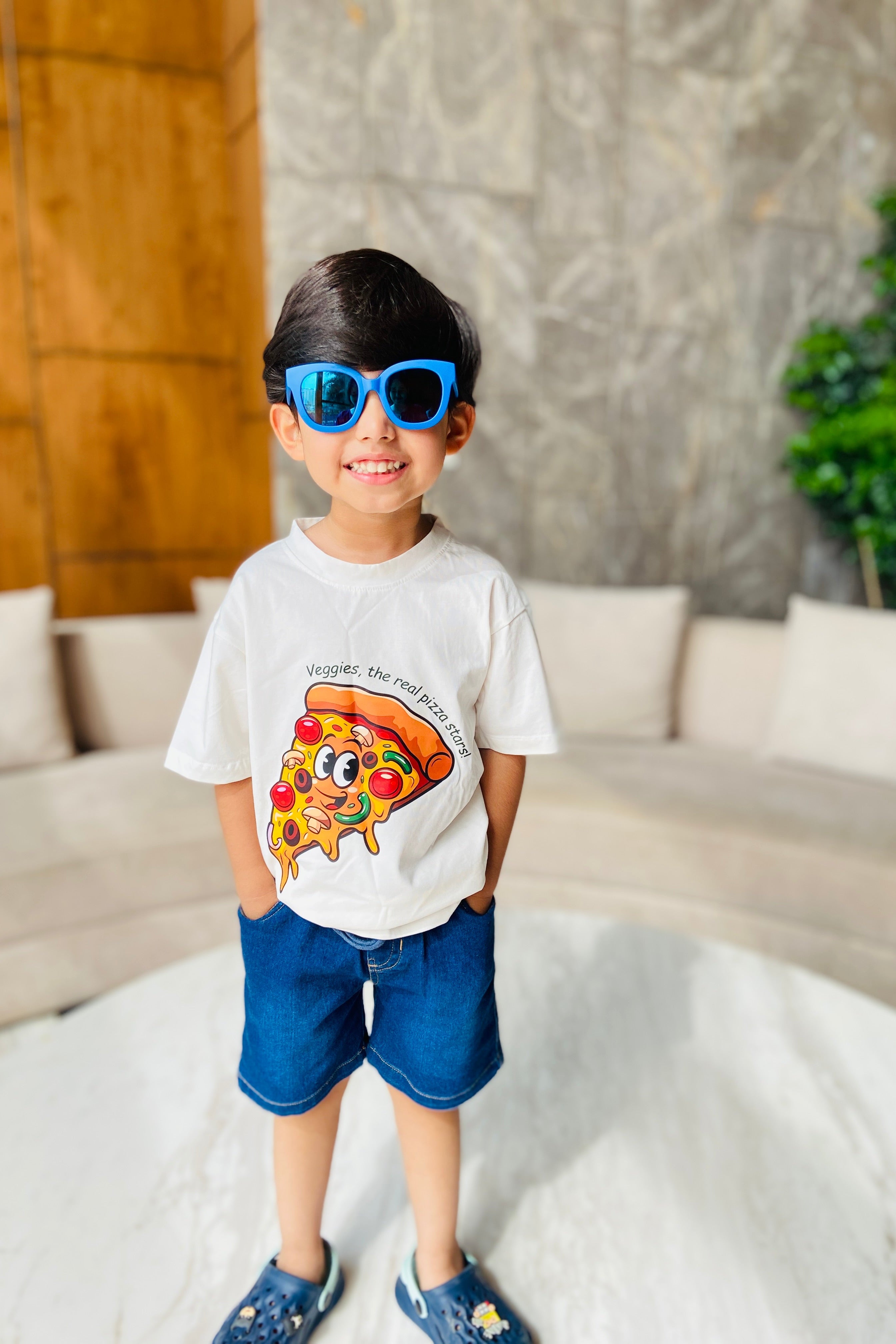 Child wearing a white t-shirt with a colorful design of Pizza print and blue shorts, standing in a room with a couch and plant.