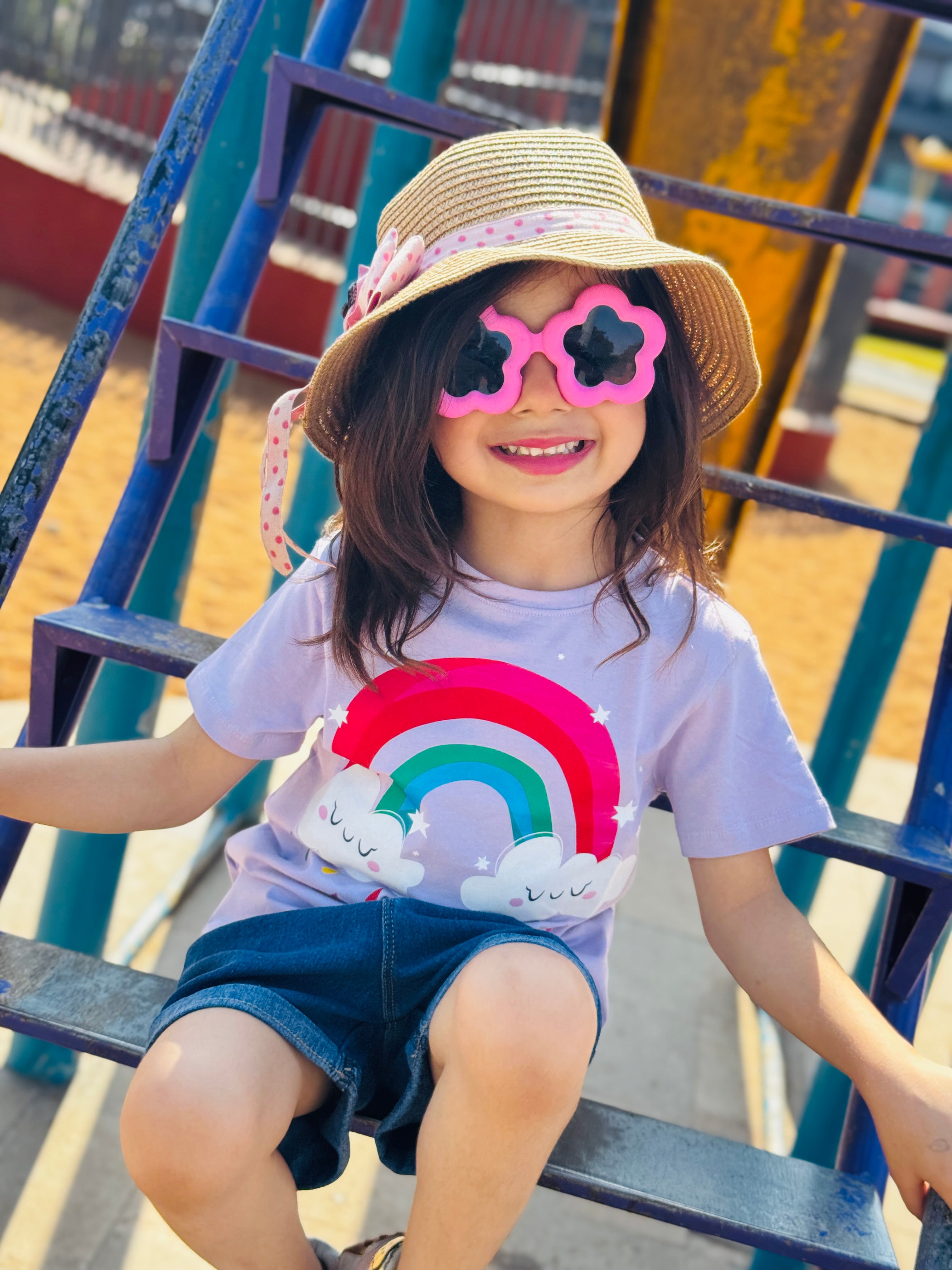 Child wearing a rainbow t-shirt and pink sunglasses sitting on a playground slide.
