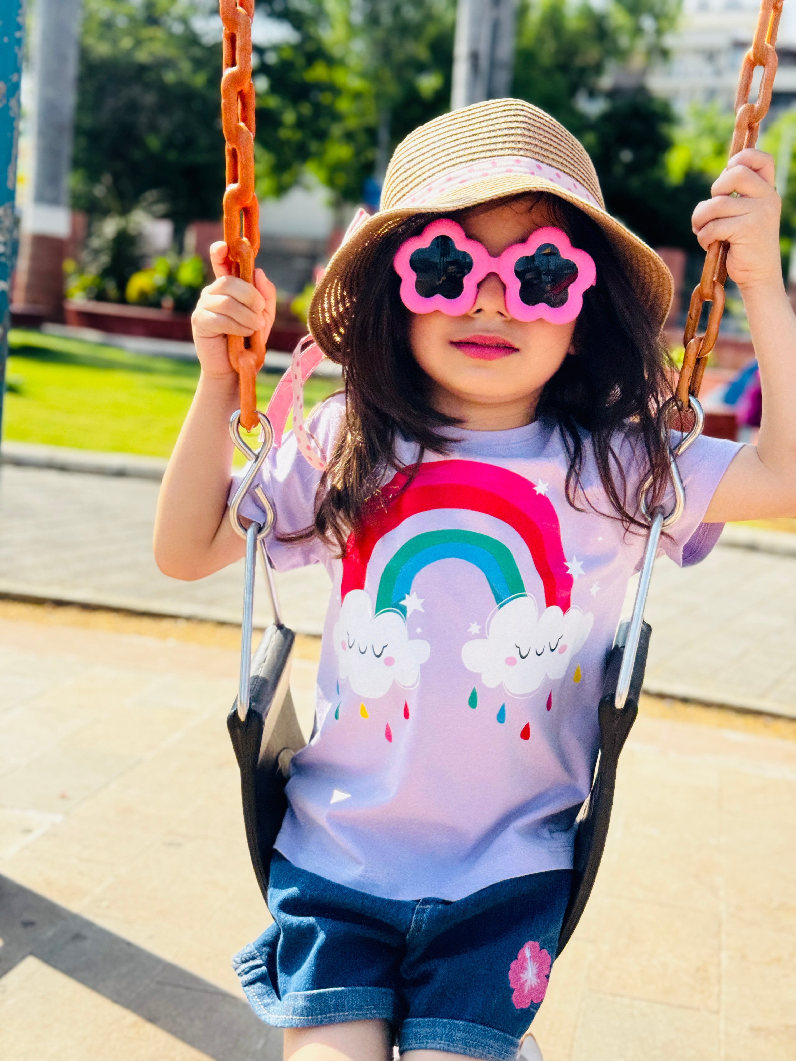 Child on a swing wearing a rainbow-themed shirt and pink sunglasses with black flower lenses.