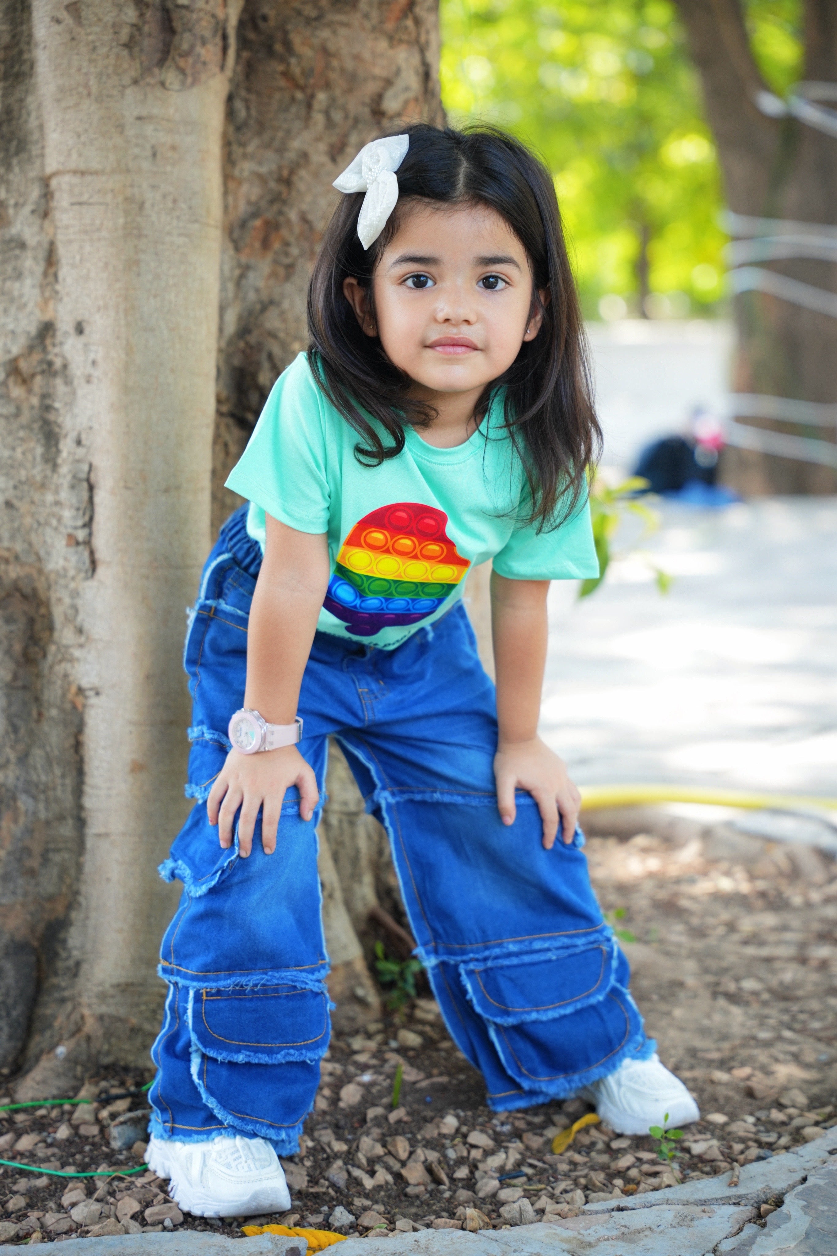 Child wearing a colorful shirt and blue pants standing outdoors near a tree.