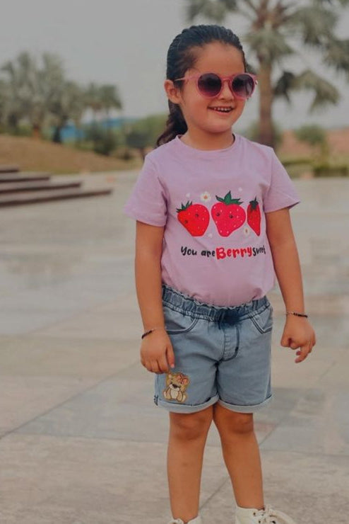Young girl wearing a lavender t-shirt with red berries and denim shorts in an outdoor setting.