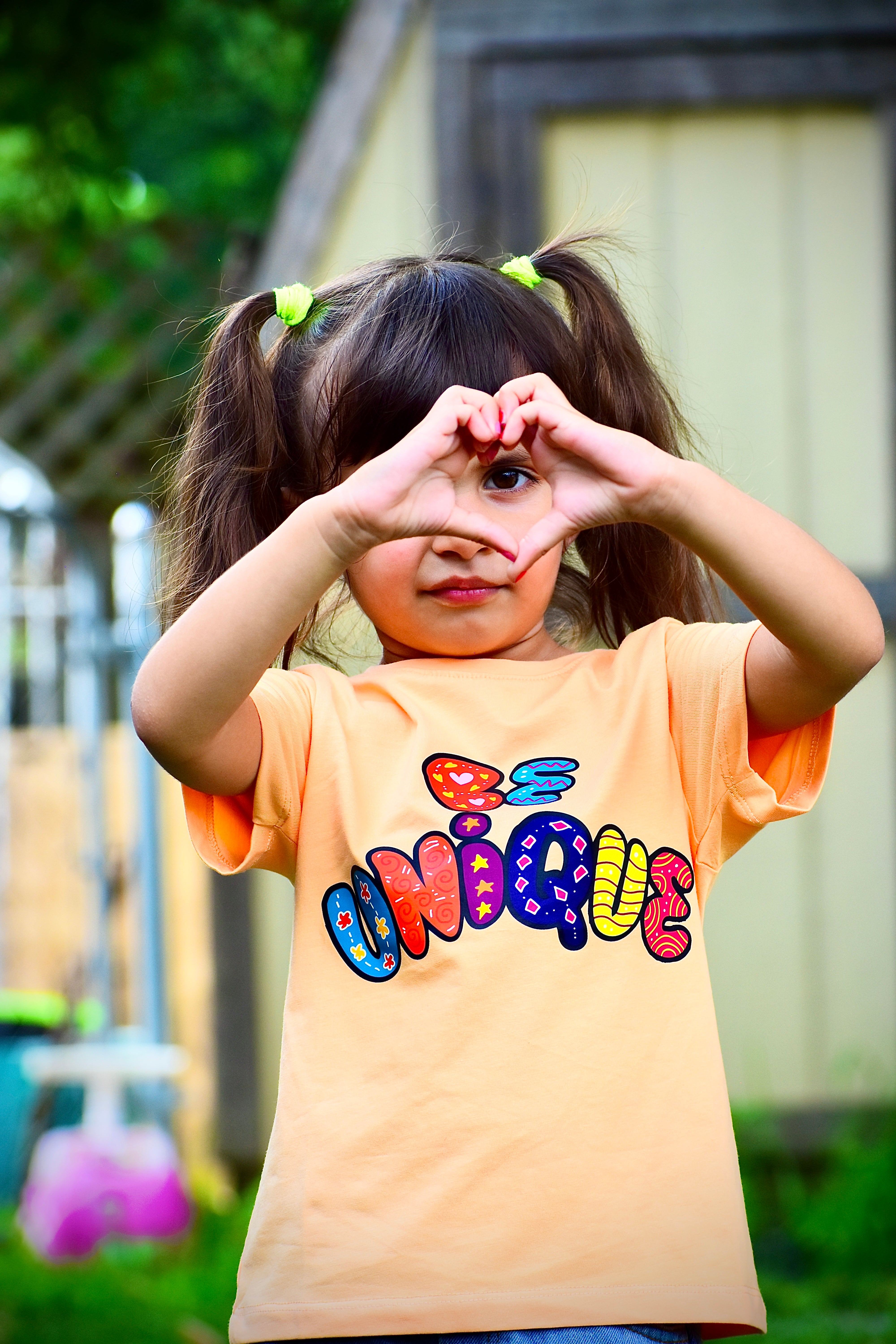 Child wearing a peach t-shirt with colorful text, making a heart shape with her hands outdoors.