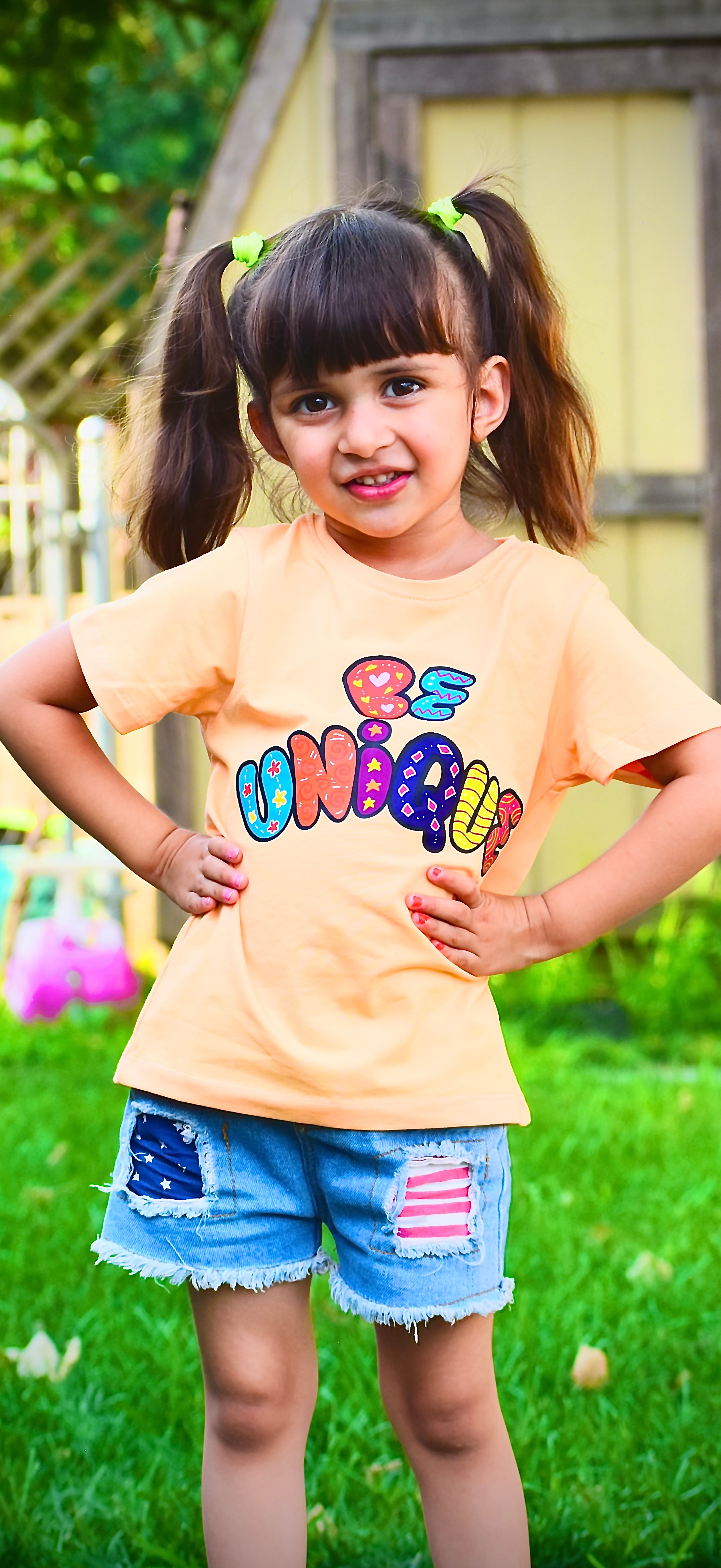 Young girl wearing a yellow t-shirt with 'Be Unique' text, standing outdoors on grass.