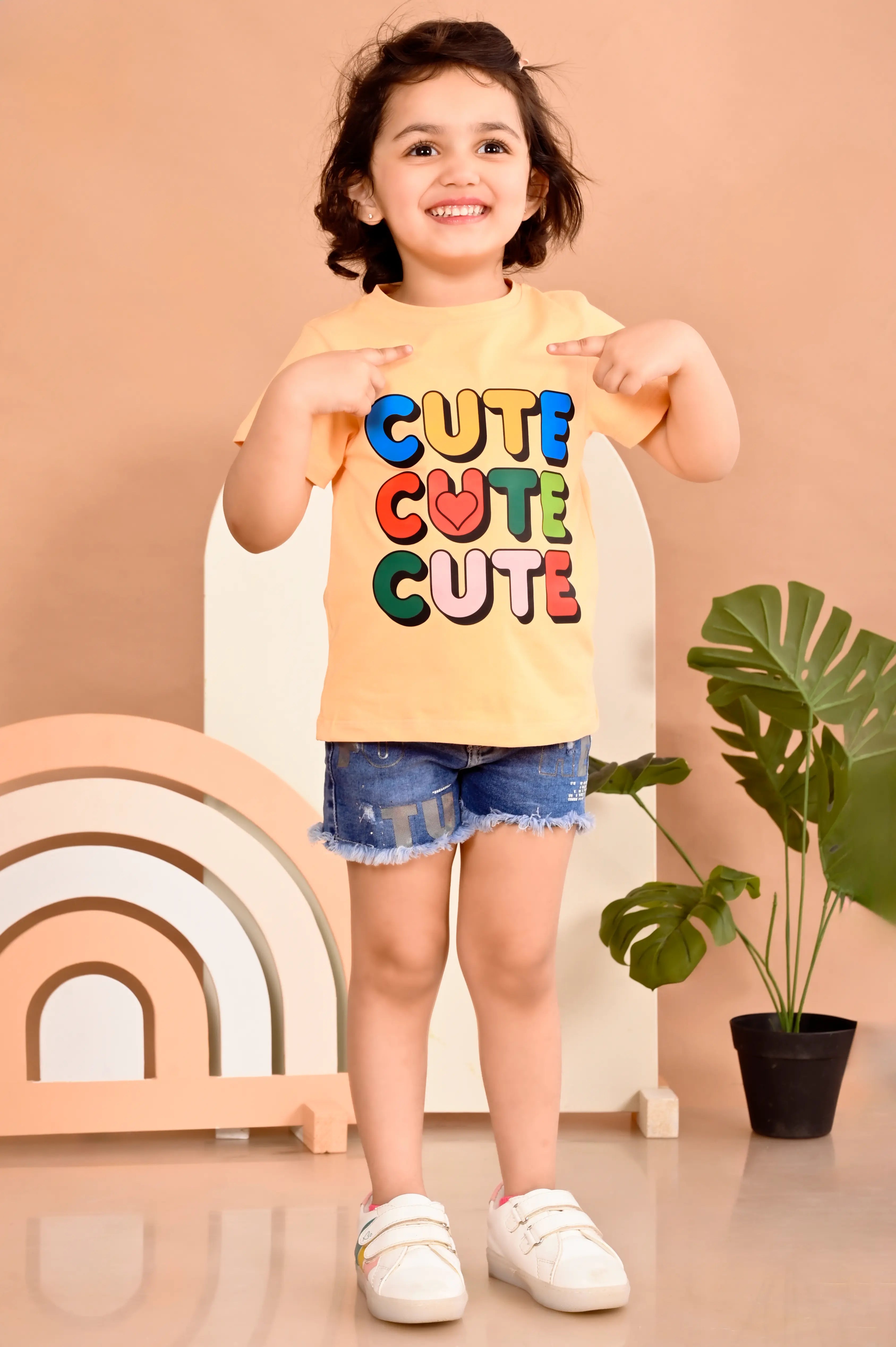 Child wearing a 'CUTE CUTE CUTE' shirt in a room with a plant and rainbow decor.