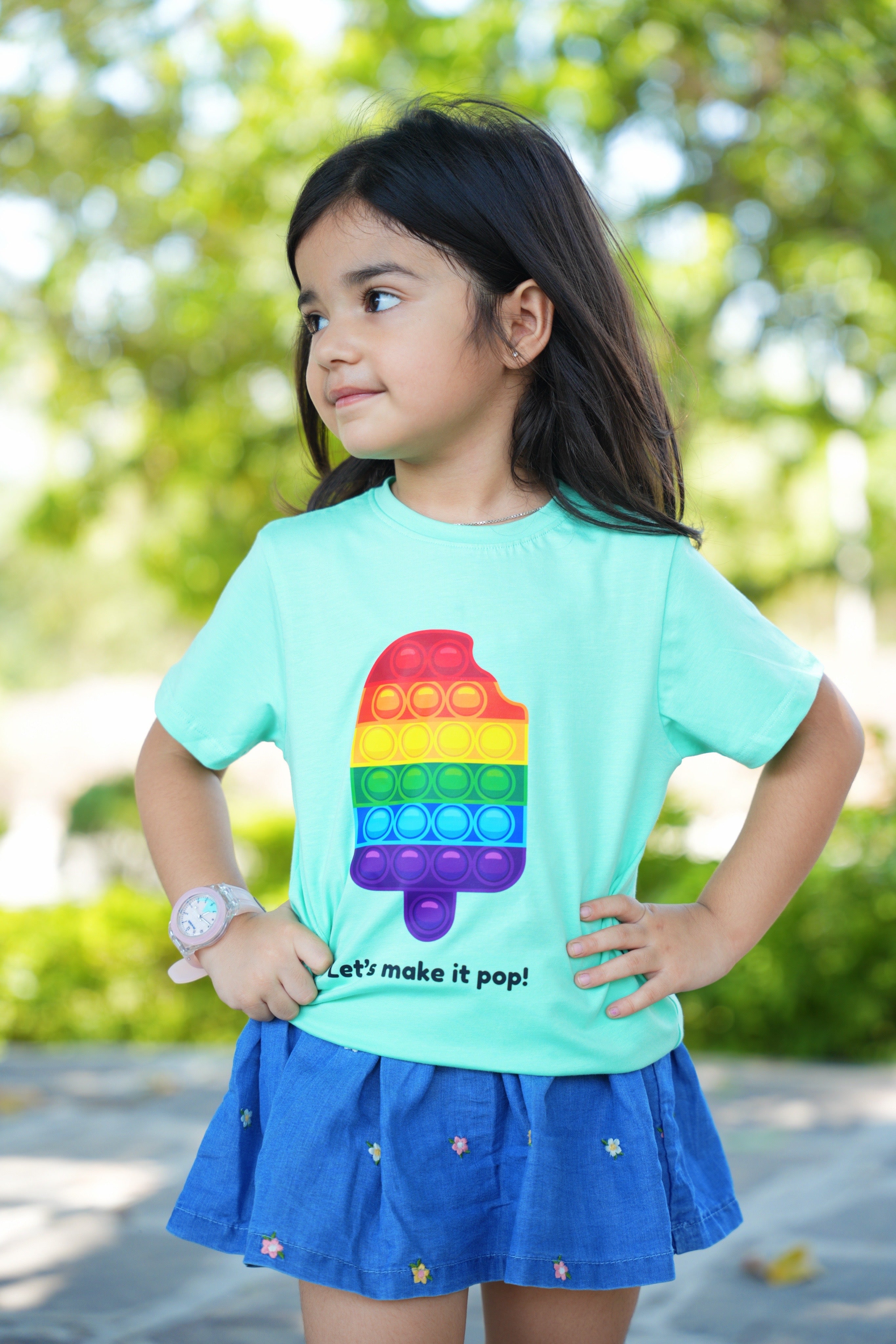 Child wearing a mint green t-shirt with a colorful pop-it design outdoors.