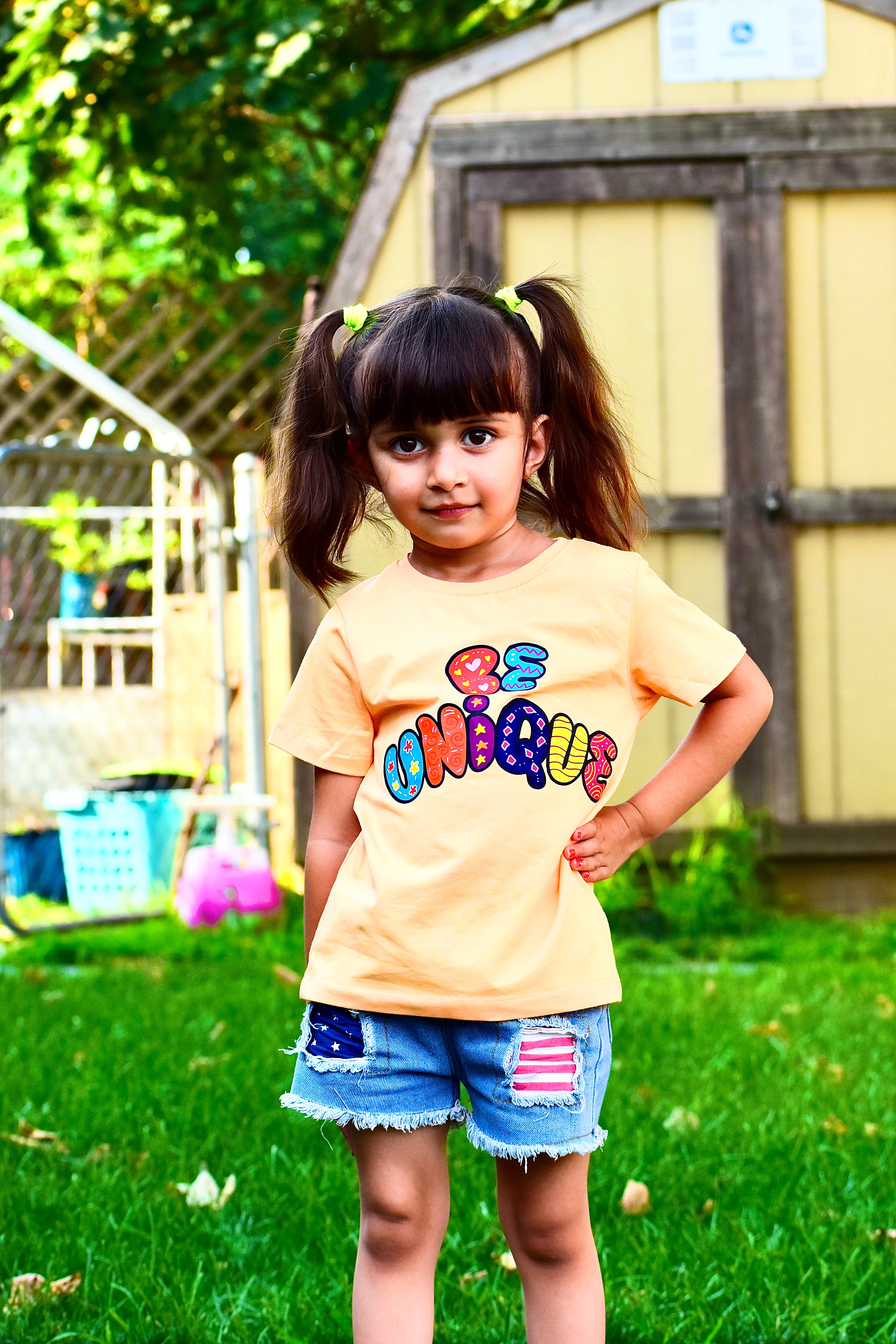 Young girl wearing a peach shirt with colorful text, standing in a backyard.